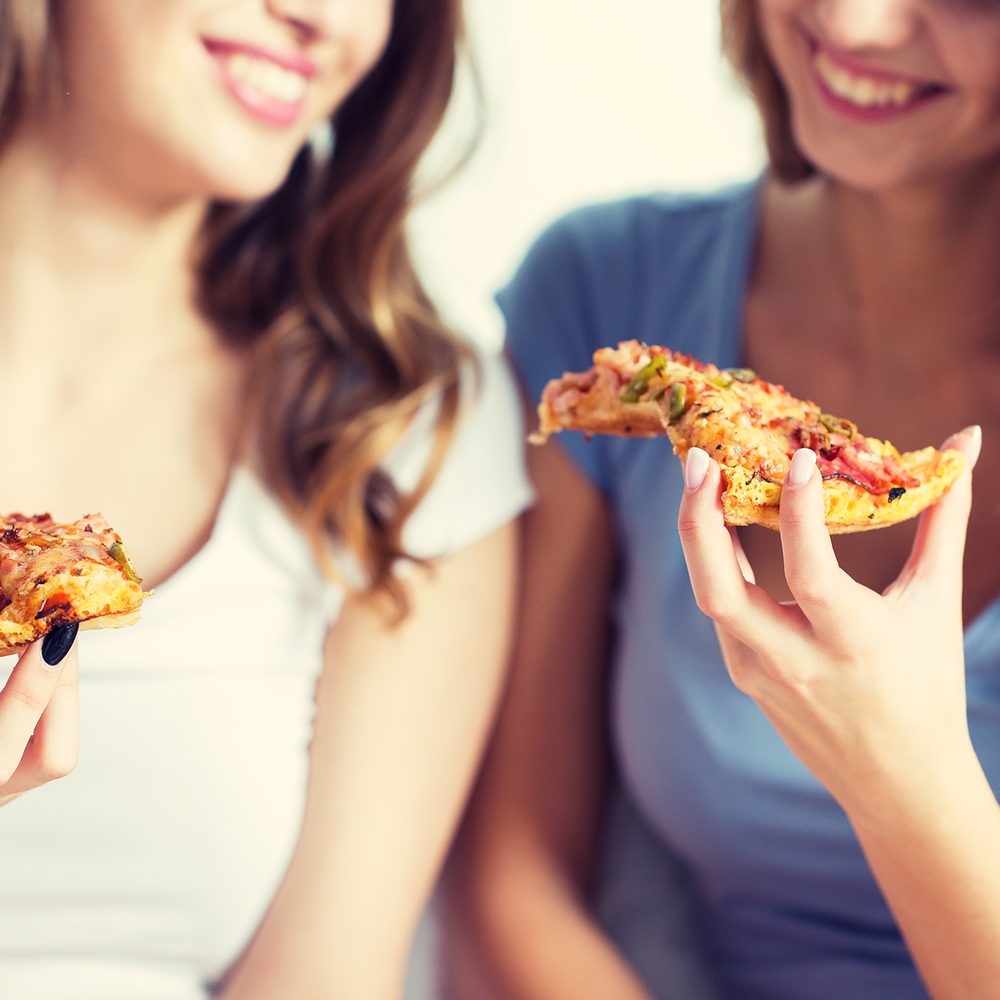 Two women smiling and sharing pizza together at a casual drop-in gathering.