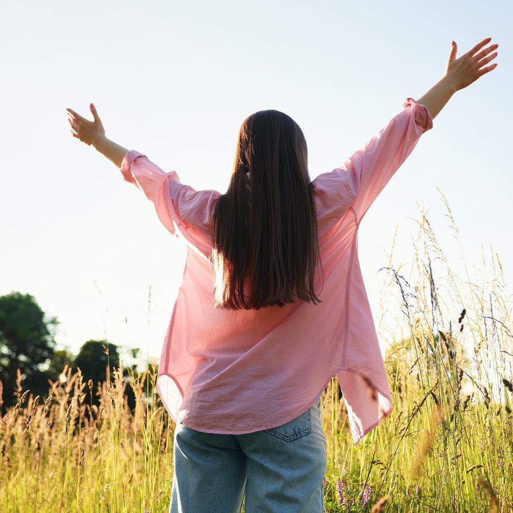 A woman standing in a sunlit field with her arms open, seen from behind. No face shown.